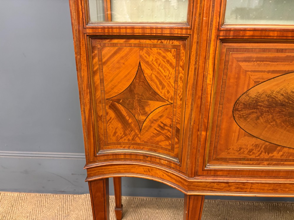 An Edwardian inlaid satinwood display cabinet featuring a central glazed door with an oval wood frame, fabric-lined shelves, and tapered square legs. View VI