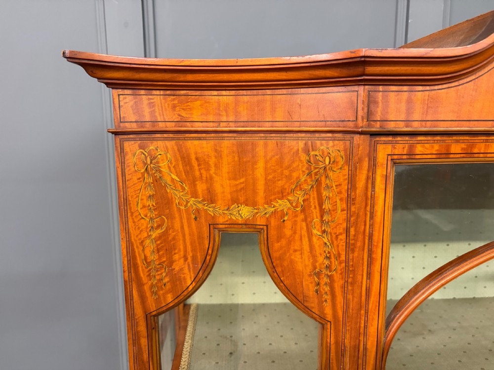 An Edwardian inlaid satinwood display cabinet featuring a central glazed door with an oval wood frame, fabric-lined shelves, and tapered square legs. View VIII
