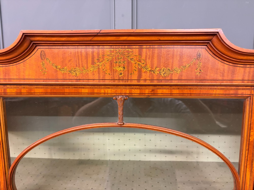 An Edwardian inlaid satinwood display cabinet featuring a central glazed door with an oval wood frame, fabric-lined shelves, and tapered square legs. View IX