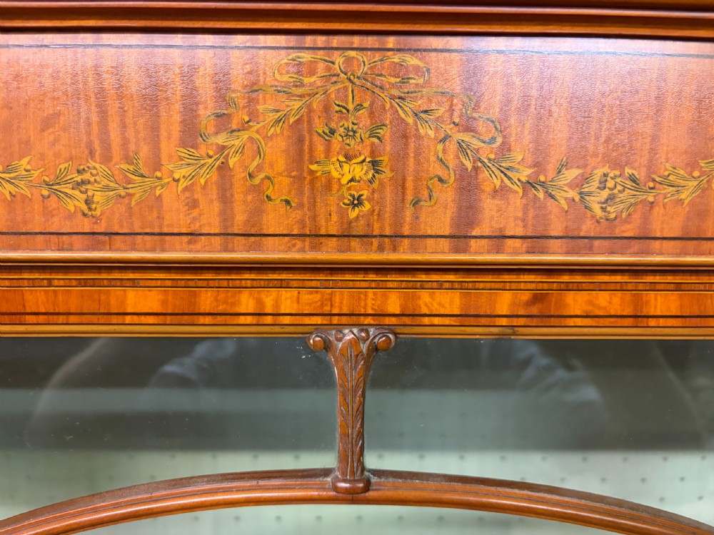 An Edwardian inlaid satinwood display cabinet featuring a central glazed door with an oval wood frame, fabric-lined shelves, and tapered square legs. View X