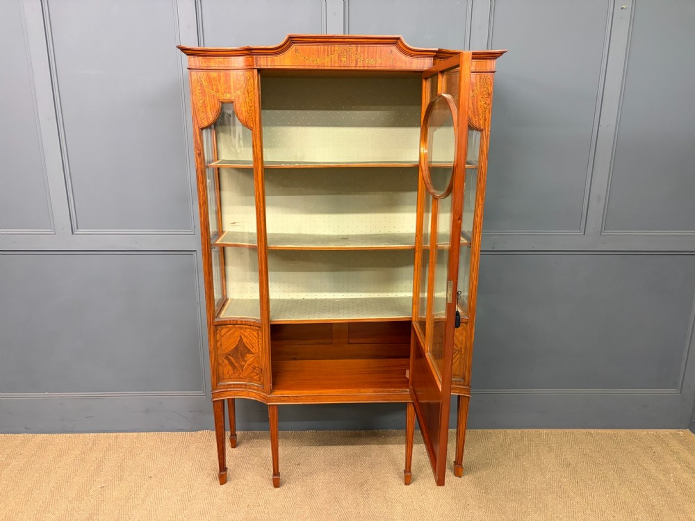 An Edwardian inlaid satinwood display cabinet featuring a central glazed door with an oval wood frame, fabric-lined shelves, and tapered square legs. View XI
