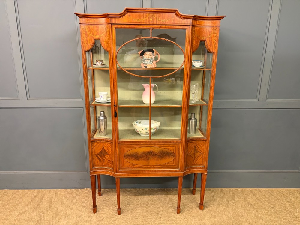 An Edwardian inlaid satinwood display cabinet featuring a central glazed door with an oval wood frame, fabric-lined shelves, and tapered square legs. View XII