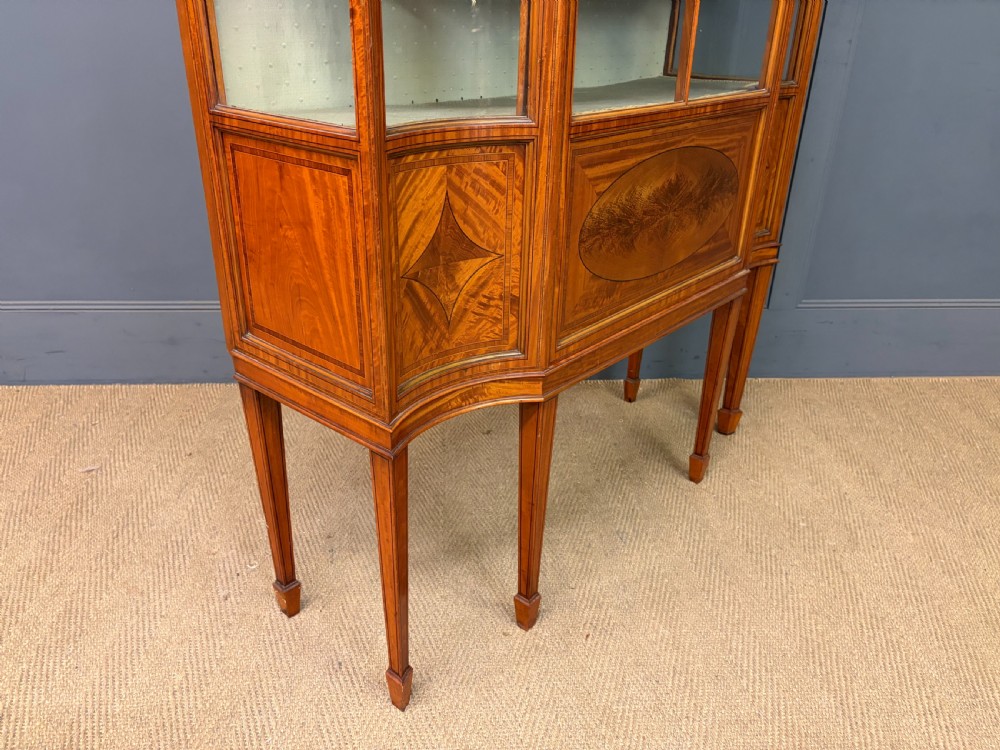 An Edwardian inlaid satinwood display cabinet featuring a central glazed door with an oval wood frame, fabric-lined shelves, and tapered square legs. View XIV