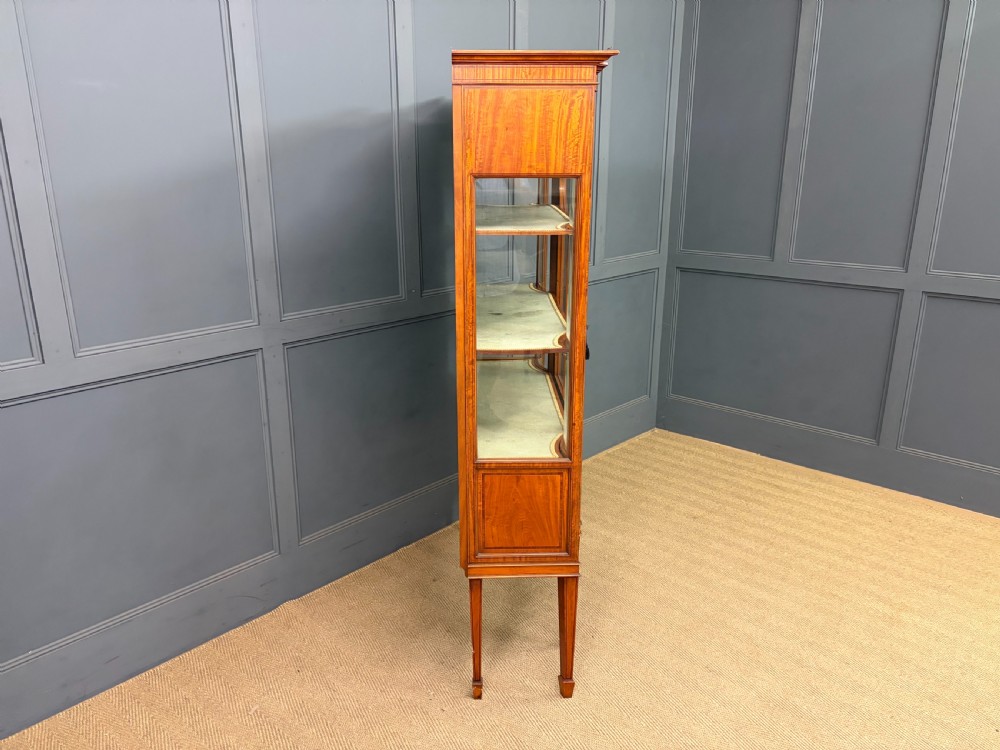 An Edwardian inlaid satinwood display cabinet featuring a central glazed door with an oval wood frame, fabric-lined shelves, and tapered square legs. View XV