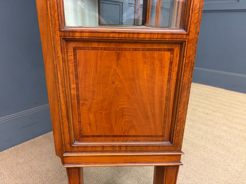 An Edwardian inlaid satinwood display cabinet featuring a central glazed door with an oval wood frame, fabric-lined shelves, and tapered square legs. View XVI