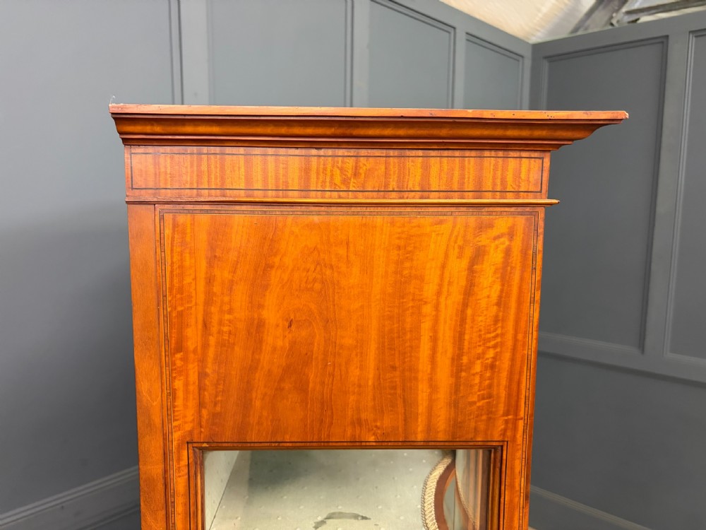 An Edwardian inlaid satinwood display cabinet featuring a central glazed door with an oval wood frame, fabric-lined shelves, and tapered square legs. View XVII