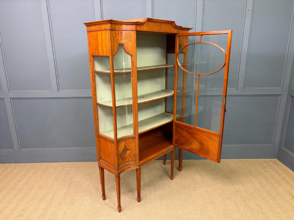 An Edwardian inlaid satinwood display cabinet featuring a central glazed door with an oval wood frame, fabric-lined shelves, and tapered square legs. View XVIII