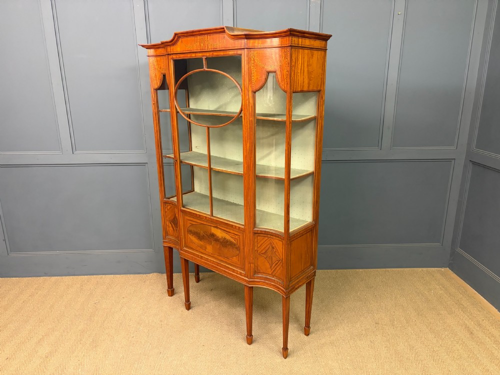 An Edwardian inlaid satinwood display cabinet featuring a central glazed door with an oval wood frame, fabric-lined shelves, and tapered square legs. View XIX