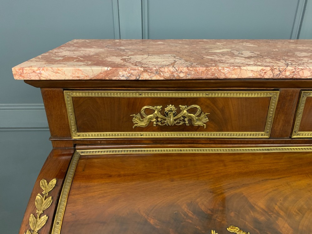 A nineteenth-century French Empire flame mahogany cylinder desk featuring a pink marble top, ornate gilt bronze mounts, and carved winged lion feet. View IV