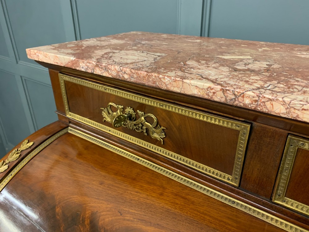 A nineteenth-century French Empire flame mahogany cylinder desk featuring a pink marble top, ornate gilt bronze mounts, and carved winged lion feet. View V