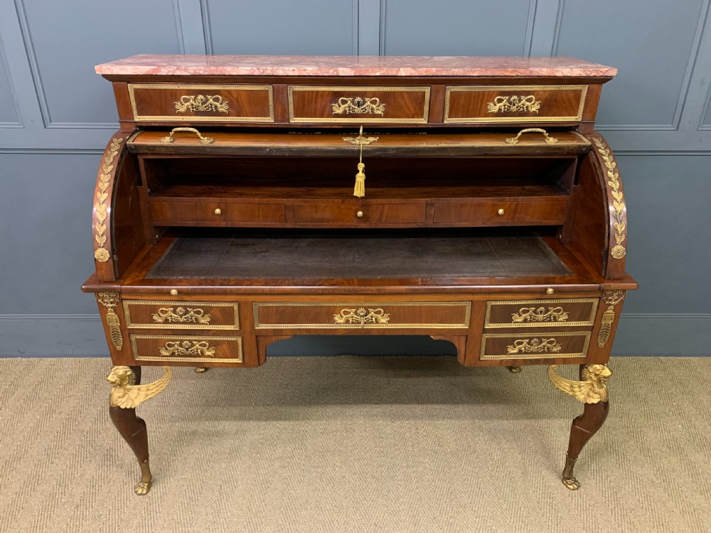 A nineteenth-century French Empire flame mahogany cylinder desk featuring a pink marble top, ornate gilt bronze mounts, and carved winged lion feet. View XVI