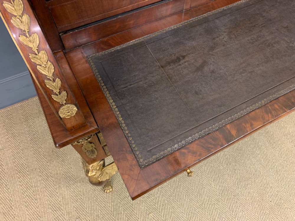 A nineteenth-century French Empire flame mahogany cylinder desk featuring a pink marble top, ornate gilt bronze mounts, and carved winged lion feet. View XVIII