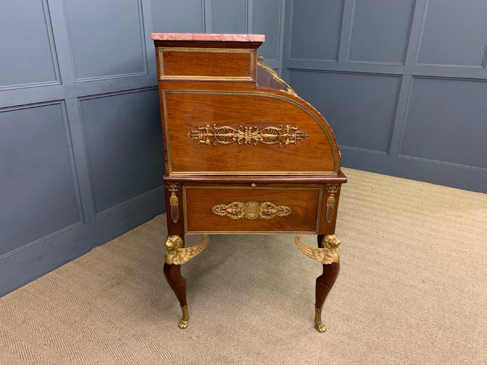 A nineteenth-century French Empire flame mahogany cylinder desk featuring a pink marble top, ornate gilt bronze mounts, and carved winged lion feet. View XIX
