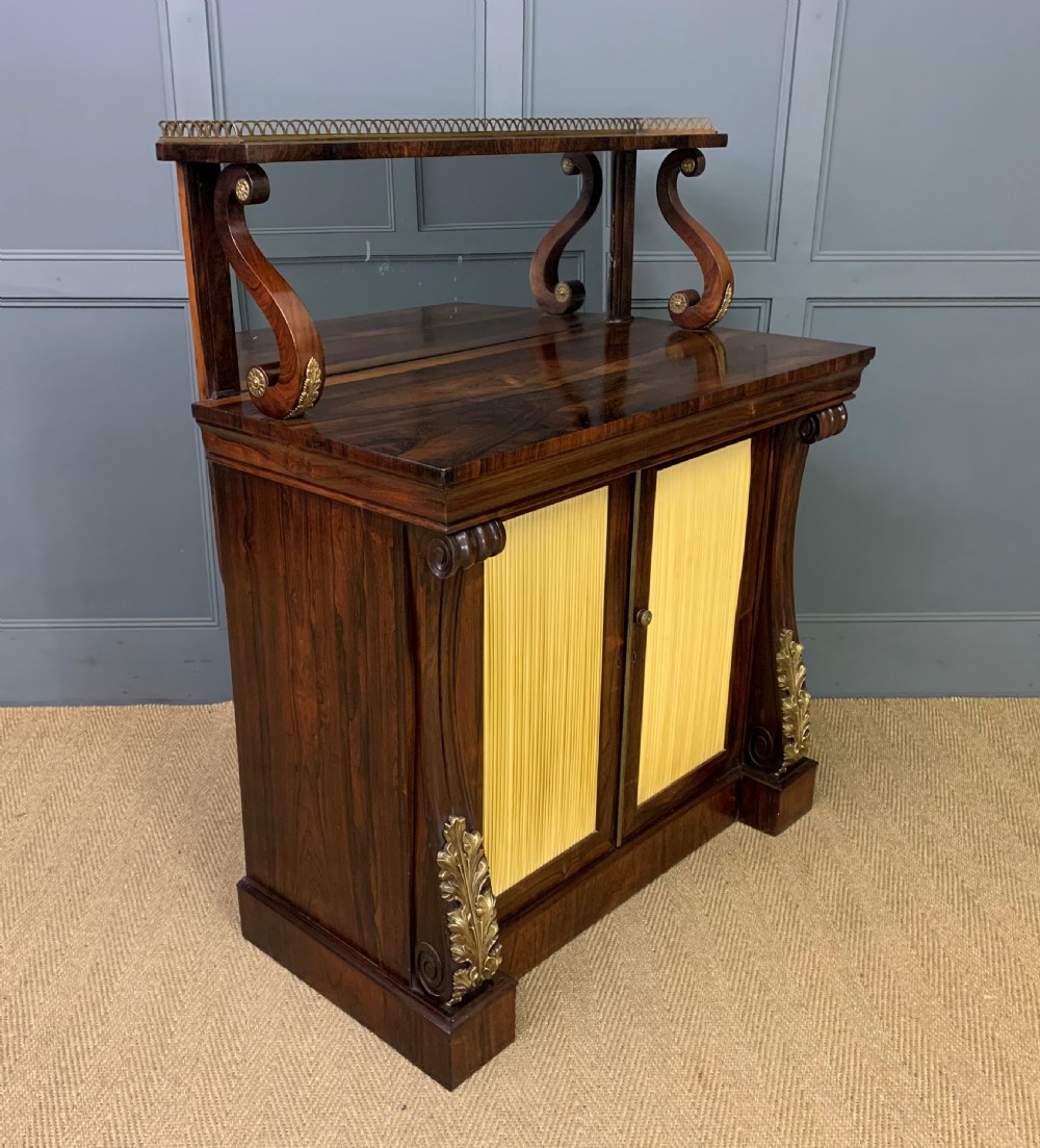 A Regency rosewood chiffonier cabinet featuring a top shelf with a brass gallery on S-scroll supports over a two-door cupboard with pleated yellow fabric panels.