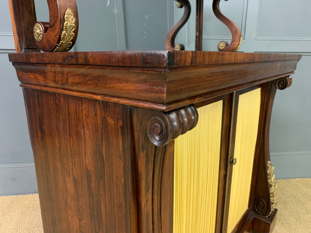 A Regency rosewood chiffonier cabinet featuring a top shelf with a brass gallery on S-scroll supports over a two-door cupboard with pleated yellow fabric panels. View X