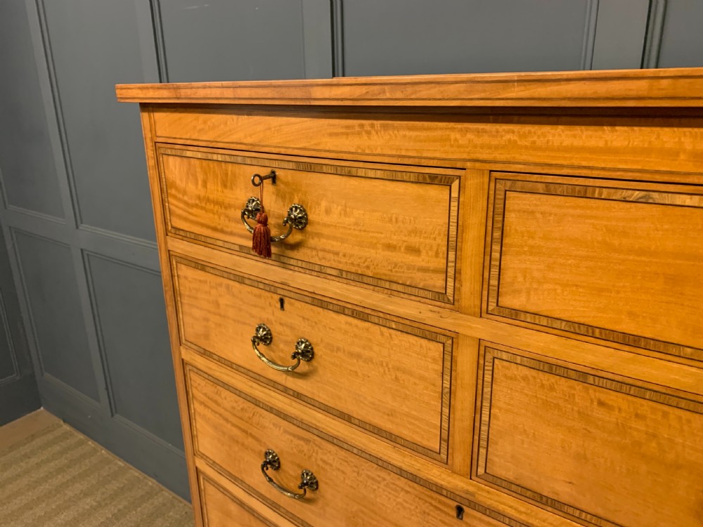 A tall satinwood chest of drawers featuring eight inlaid drawers with brass swan-neck handles and bracket feet. View V