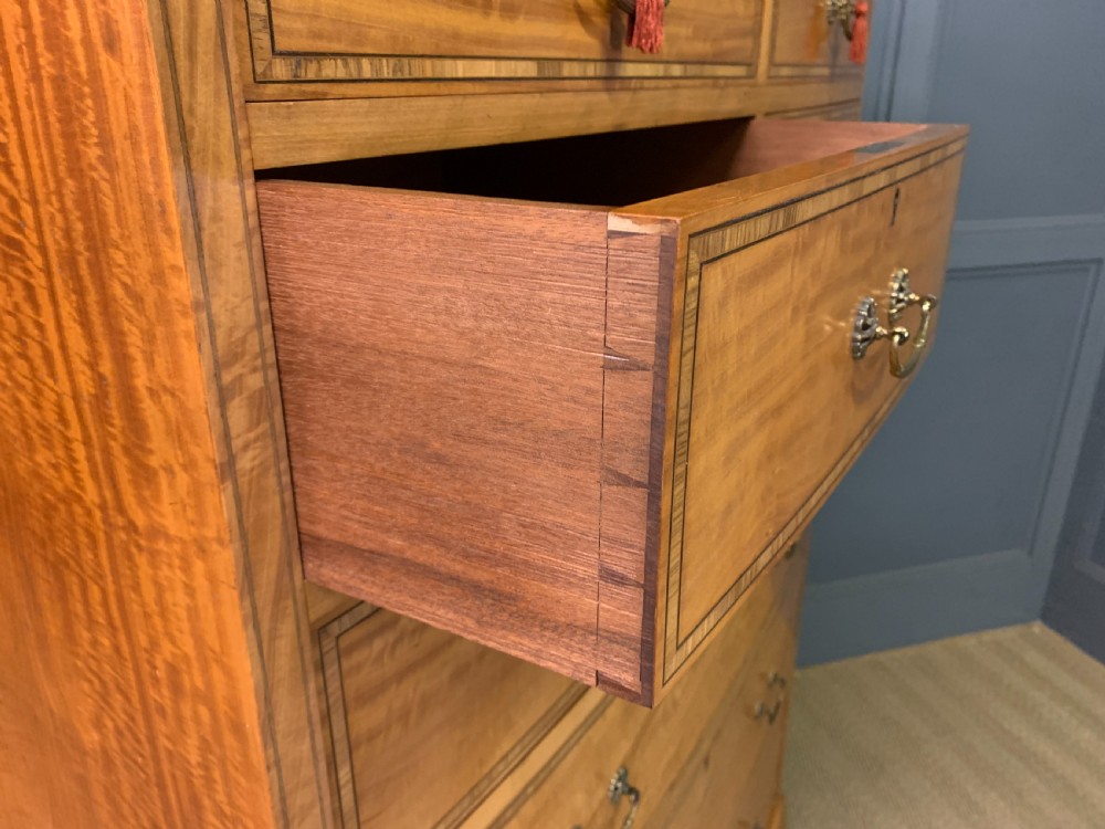 A tall satinwood chest of drawers featuring eight inlaid drawers with brass swan-neck handles and bracket feet. View XVI