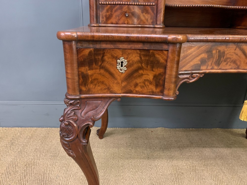 A flame mahogany writing desk featuring carved cabriole legs, a raised superstructure with drawers and gallery shelves, and a curved front with brass keyholes. View IV