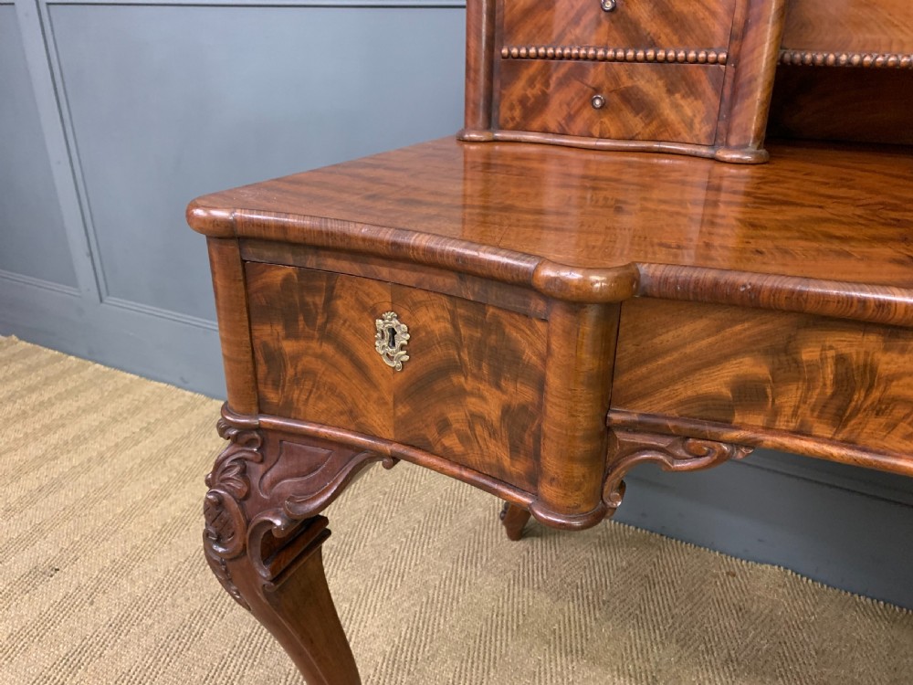 A flame mahogany writing desk featuring carved cabriole legs, a raised superstructure with drawers and gallery shelves, and a curved front with brass keyholes. View V