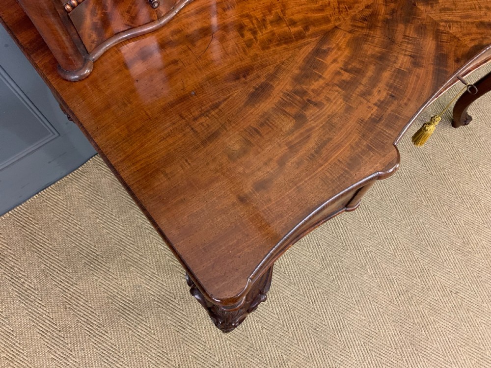 A flame mahogany writing desk featuring carved cabriole legs, a raised superstructure with drawers and gallery shelves, and a curved front with brass keyholes. View VI