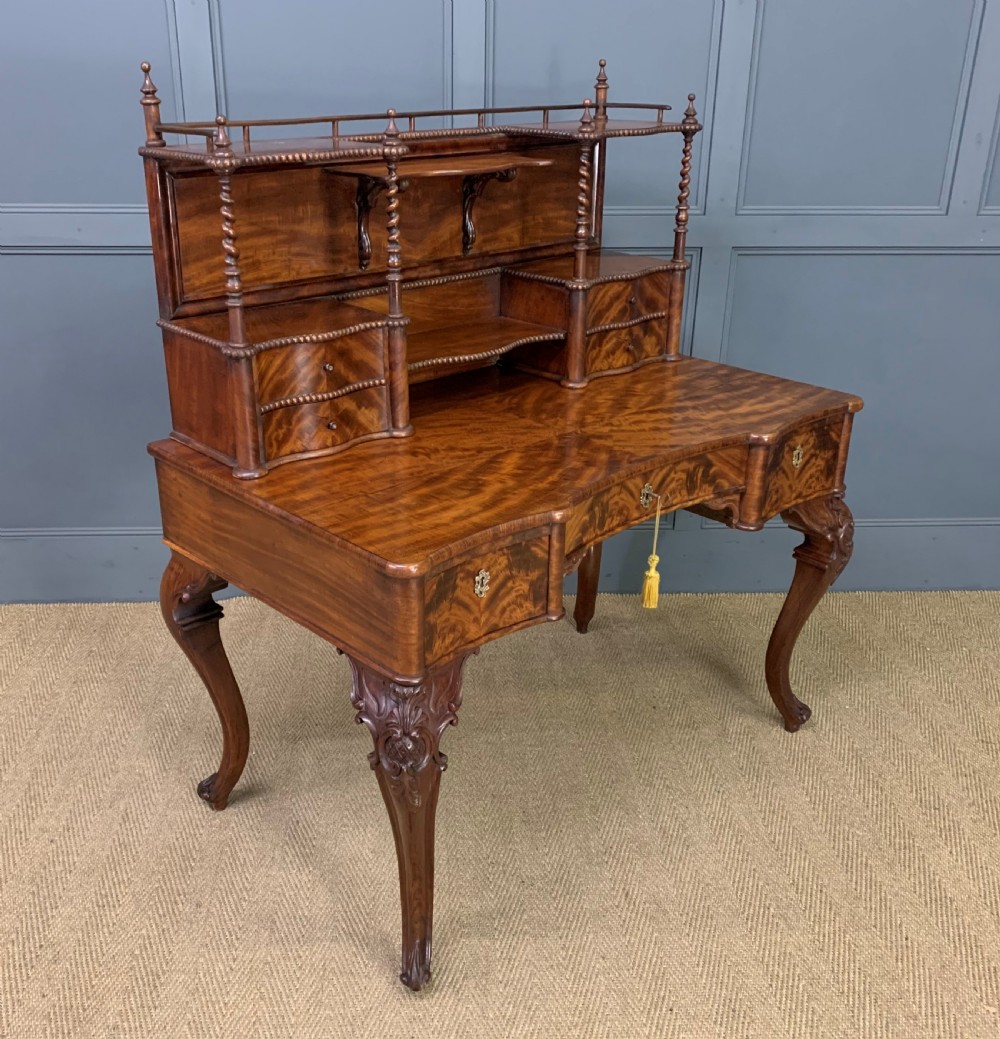 A flame mahogany writing desk featuring carved cabriole legs, a raised superstructure with drawers and gallery shelves, and a curved front with brass keyholes. View XI