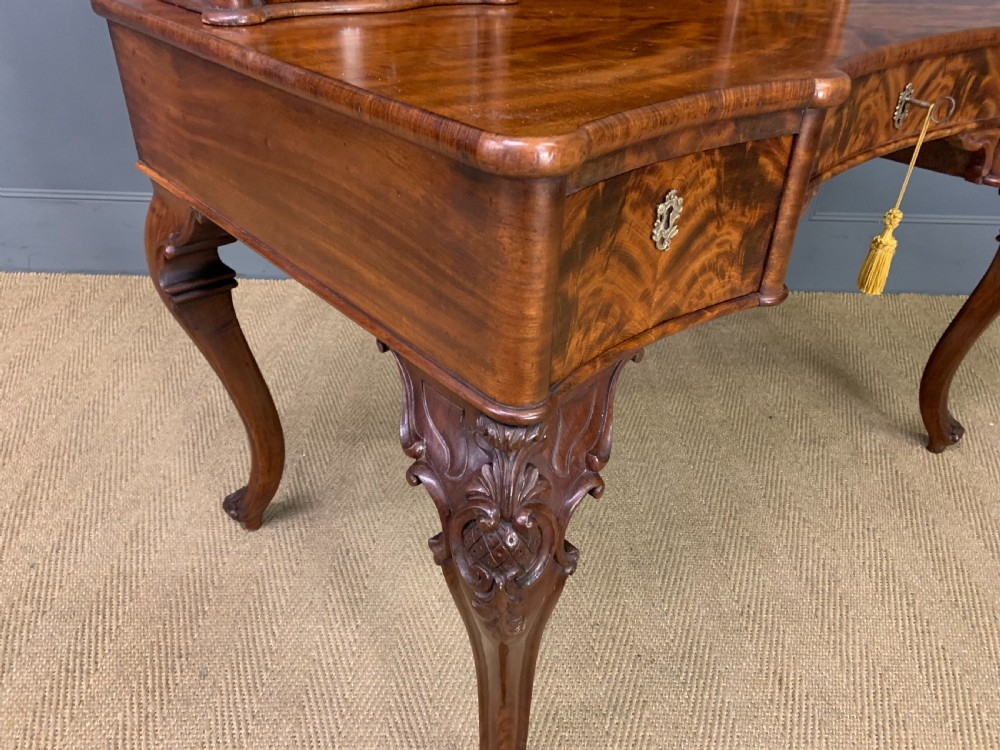 A flame mahogany writing desk featuring carved cabriole legs, a raised superstructure with drawers and gallery shelves, and a curved front with brass keyholes. View XIII