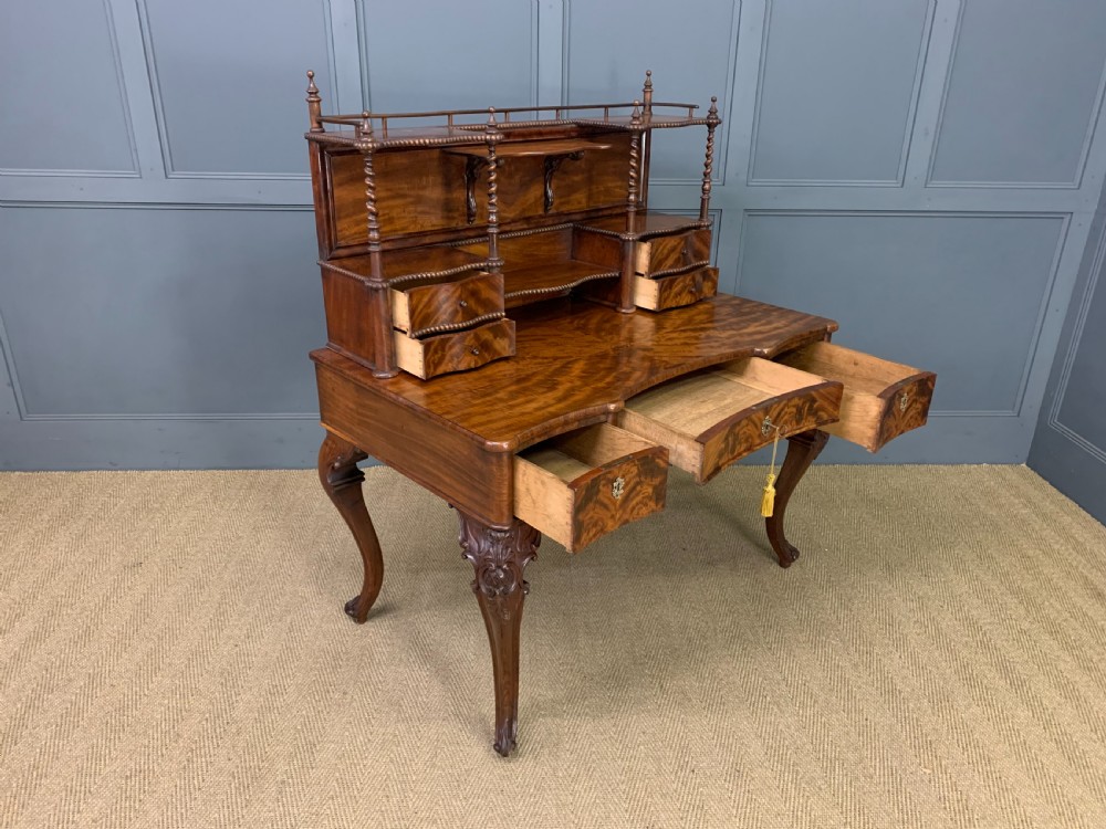 A flame mahogany writing desk featuring carved cabriole legs, a raised superstructure with drawers and gallery shelves, and a curved front with brass keyholes. View XV
