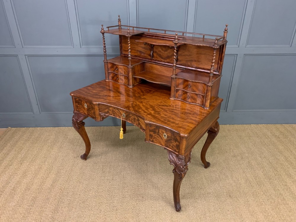 A flame mahogany writing desk featuring carved cabriole legs, a raised superstructure with drawers and gallery shelves, and a curved front with brass keyholes. View XVI