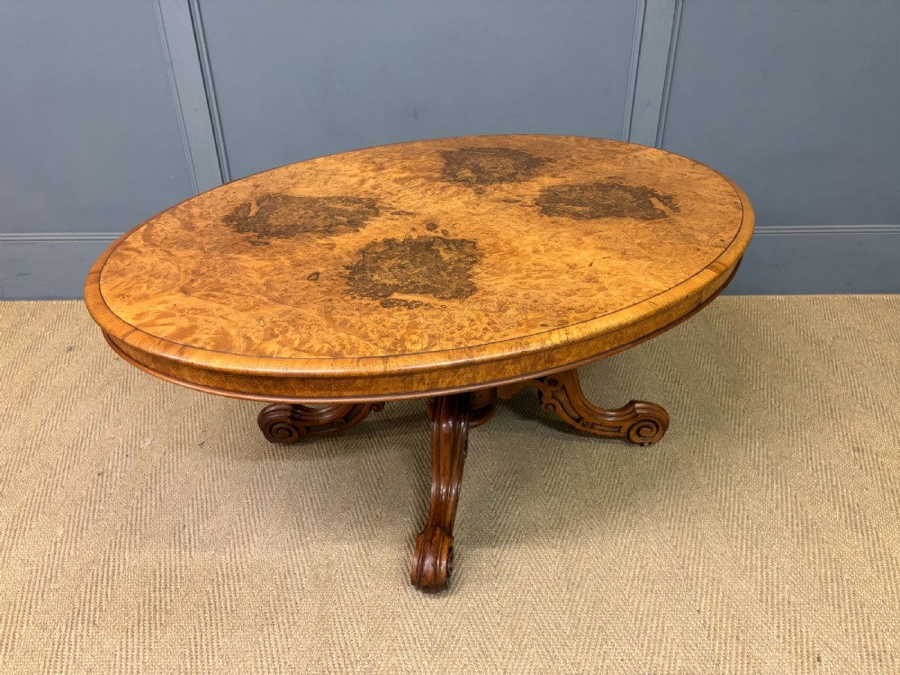 An oval Victorian coffee table crafted from figured burr walnut, featuring a carved four-legged pedestal base and scroll feet.