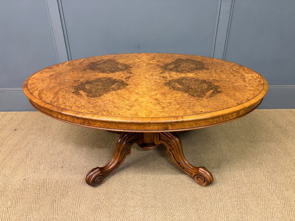 An oval Victorian coffee table crafted from figured burr walnut, featuring a carved four-legged pedestal base and scroll feet. View III