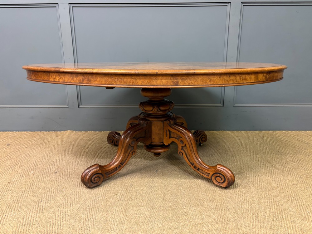 An oval Victorian coffee table crafted from figured burr walnut, featuring a carved four-legged pedestal base and scroll feet. View IV