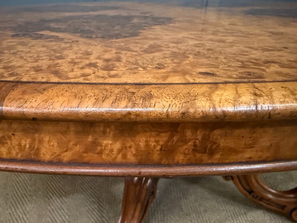 An oval Victorian coffee table crafted from figured burr walnut, featuring a carved four-legged pedestal base and scroll feet. View VI