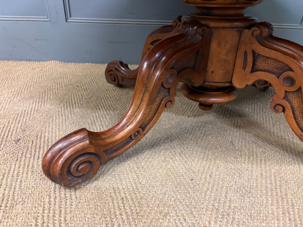 An oval Victorian coffee table crafted from figured burr walnut, featuring a carved four-legged pedestal base and scroll feet. View XIII