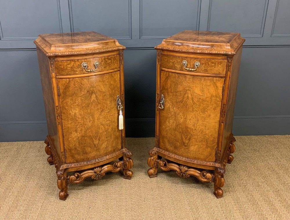A pair of tall burr walnut pedestal cupboards featuring curved front doors, single top drawers with brass drop handles, and carved scroll feet.