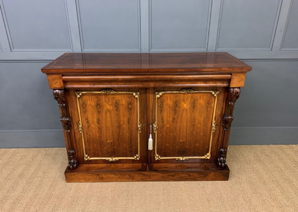 A dark Regency period rosewood side cabinet featuring two cupboard doors with gilded bronze trim and carved scroll supports on a plinth base.