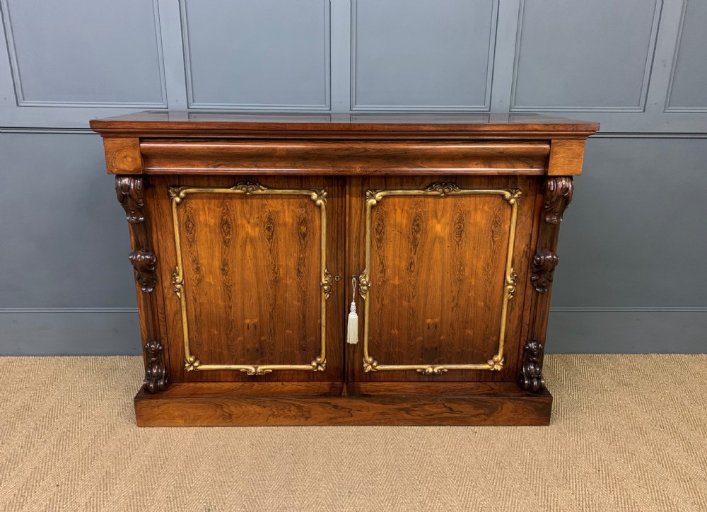 A dark Regency period rosewood side cabinet featuring two cupboard doors with gilded bronze trim and carved scroll supports on a plinth base. View III