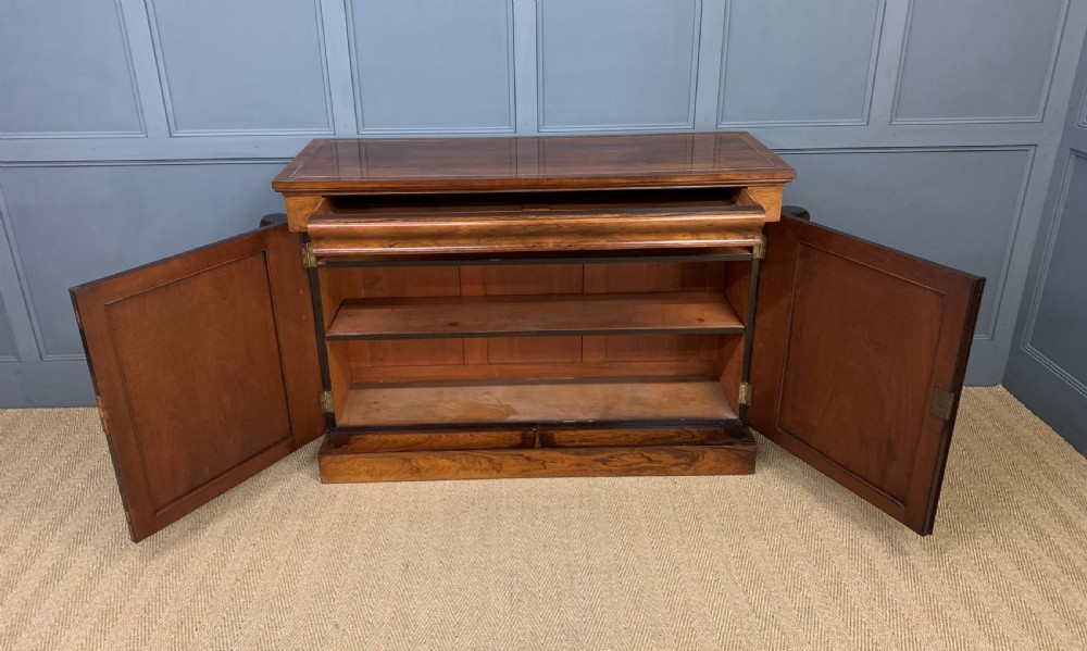 A dark Regency period rosewood side cabinet featuring two cupboard doors with gilded bronze trim and carved scroll supports on a plinth base. View IX