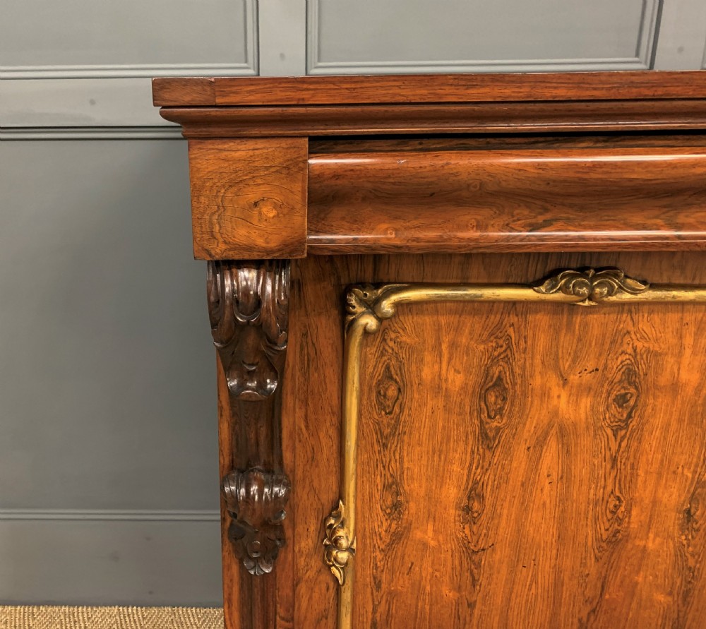 A dark Regency period rosewood side cabinet featuring two cupboard doors with gilded bronze trim and carved scroll supports on a plinth base. View X