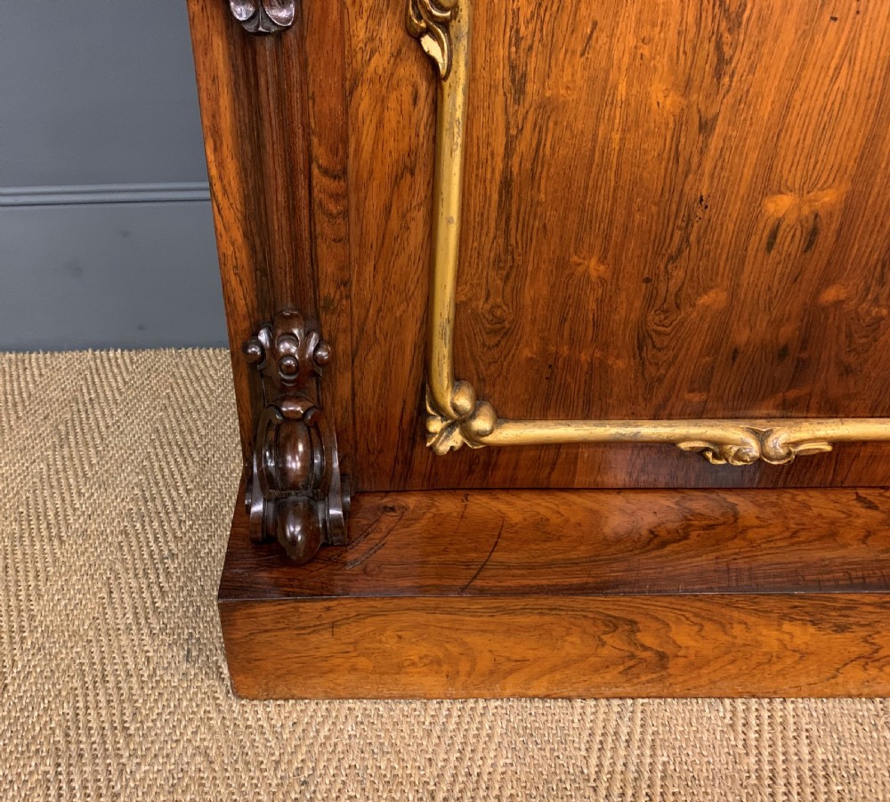 A dark Regency period rosewood side cabinet featuring two cupboard doors with gilded bronze trim and carved scroll supports on a plinth base. View XI