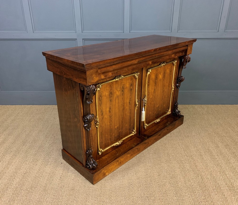 A dark Regency period rosewood side cabinet featuring two cupboard doors with gilded bronze trim and carved scroll supports on a plinth base. View XIV