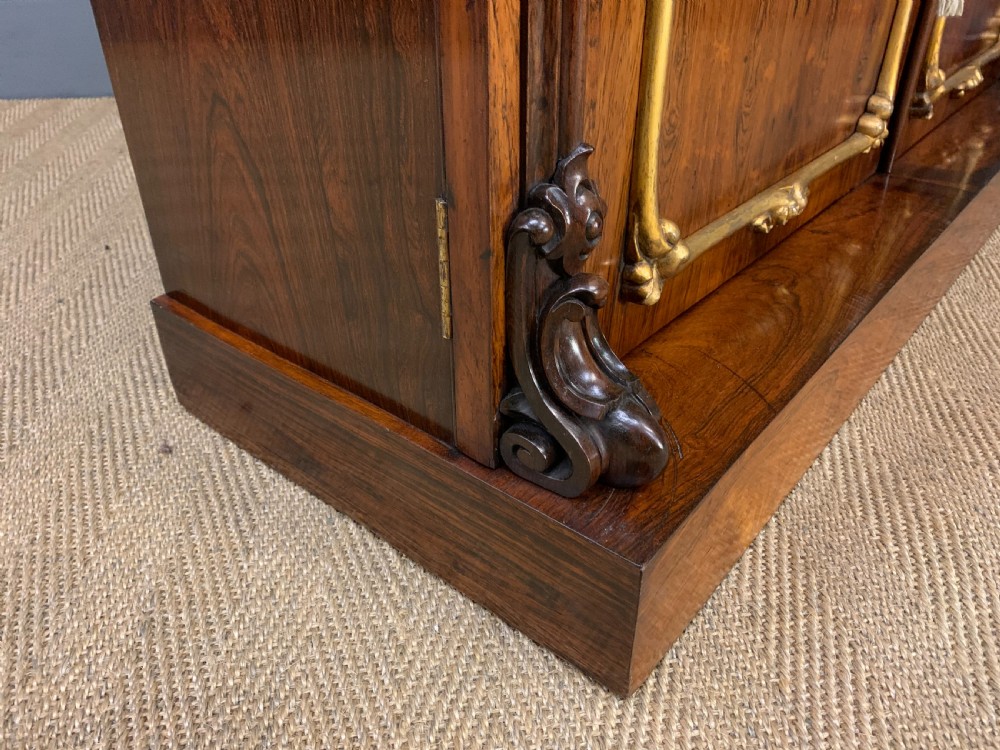 A dark Regency period rosewood side cabinet featuring two cupboard doors with gilded bronze trim and carved scroll supports on a plinth base. View XVIII