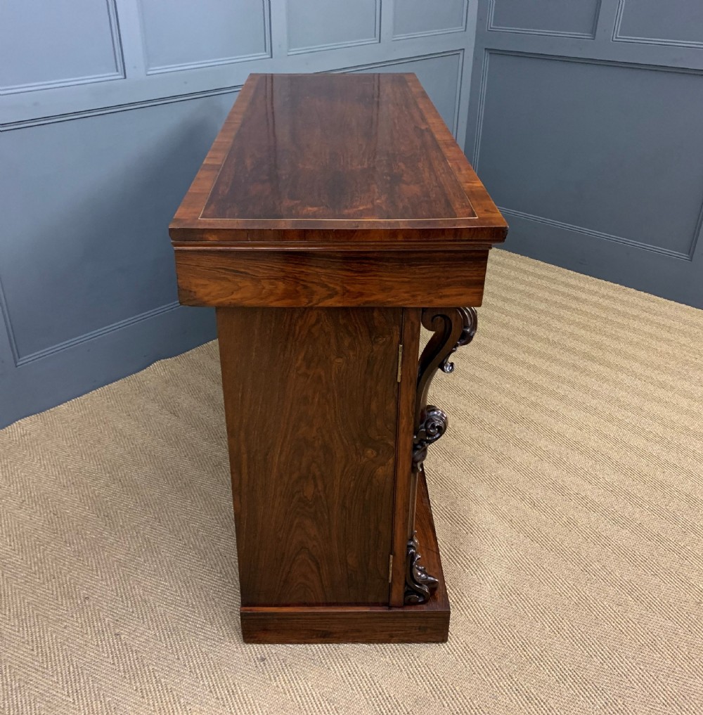 A dark Regency period rosewood side cabinet featuring two cupboard doors with gilded bronze trim and carved scroll supports on a plinth base. View XIX