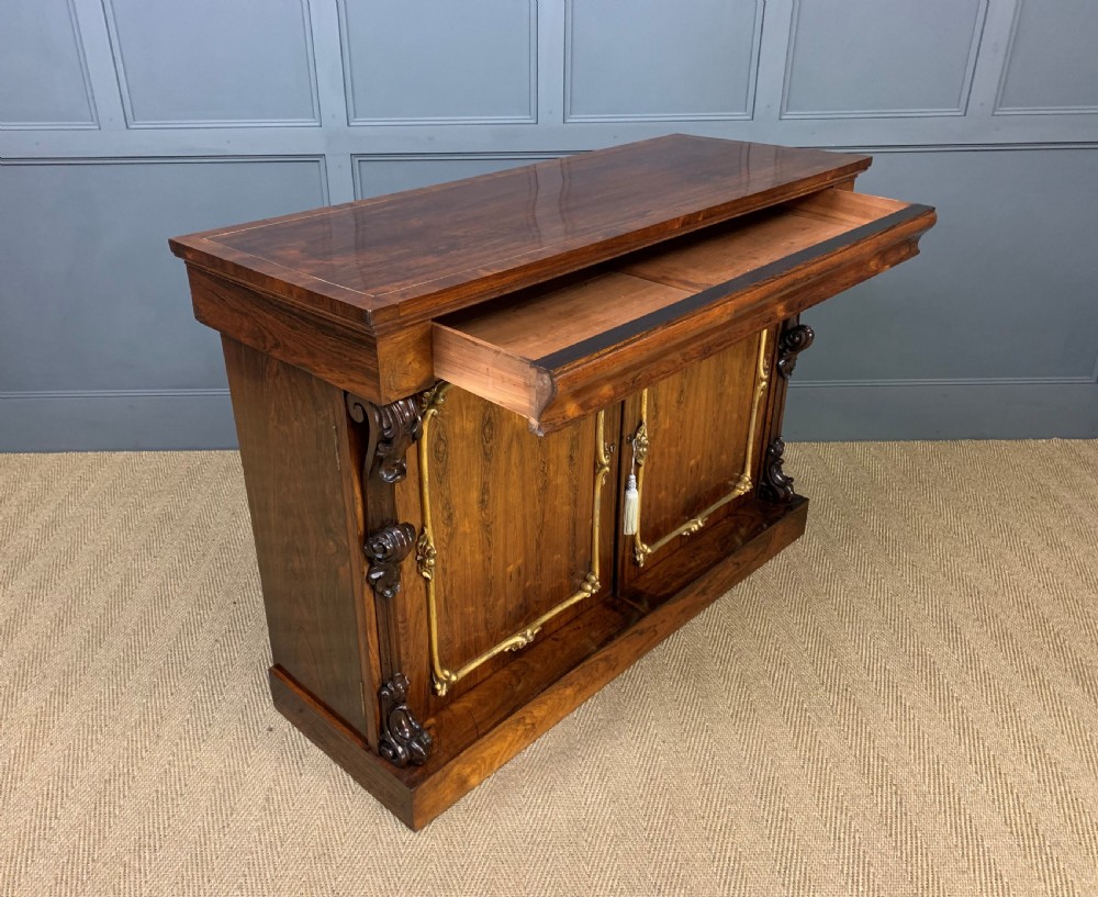 A dark Regency period rosewood side cabinet featuring two cupboard doors with gilded bronze trim and carved scroll supports on a plinth base. View XX