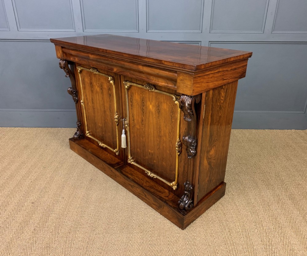 A dark Regency period rosewood side cabinet featuring two cupboard doors with gilded bronze trim and carved scroll supports on a plinth base. View XXI
