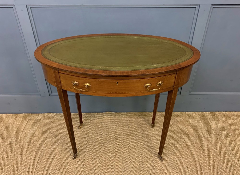 An Edwardian oval mahogany writing table featuring a green leather inset top, single frieze drawer with brass bail handles, and square tapered legs with castors.