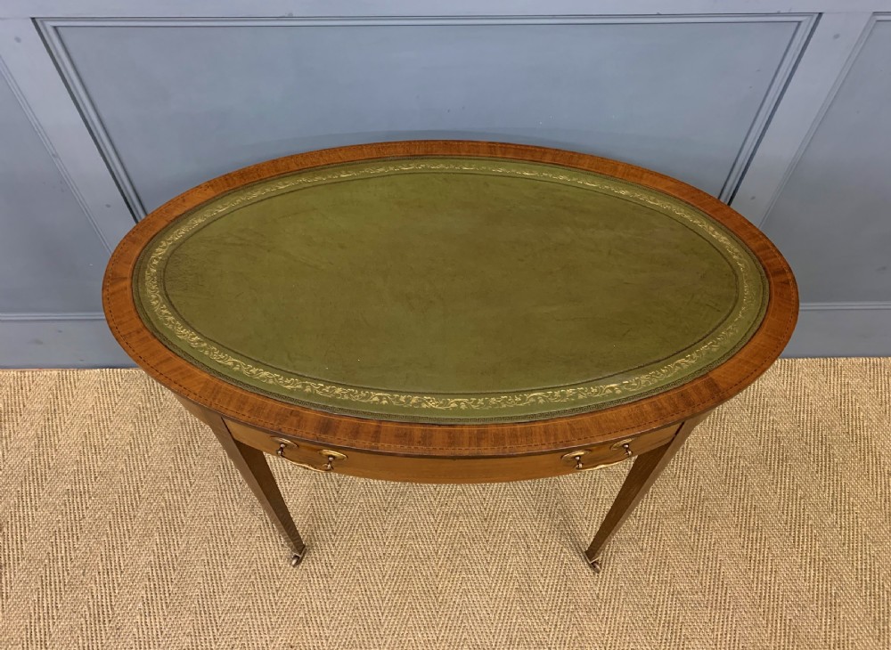 An Edwardian oval mahogany writing table featuring a green leather inset top, single frieze drawer with brass bail handles, and square tapered legs with castors. View II