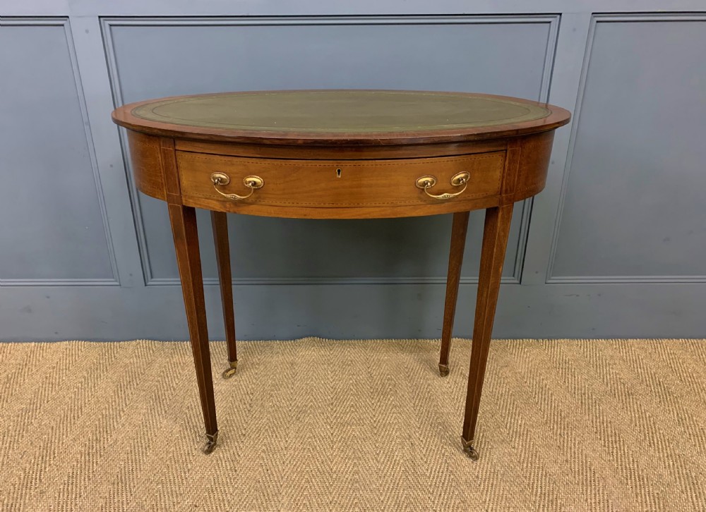 An Edwardian oval mahogany writing table featuring a green leather inset top, single frieze drawer with brass bail handles, and square tapered legs with castors. View III
