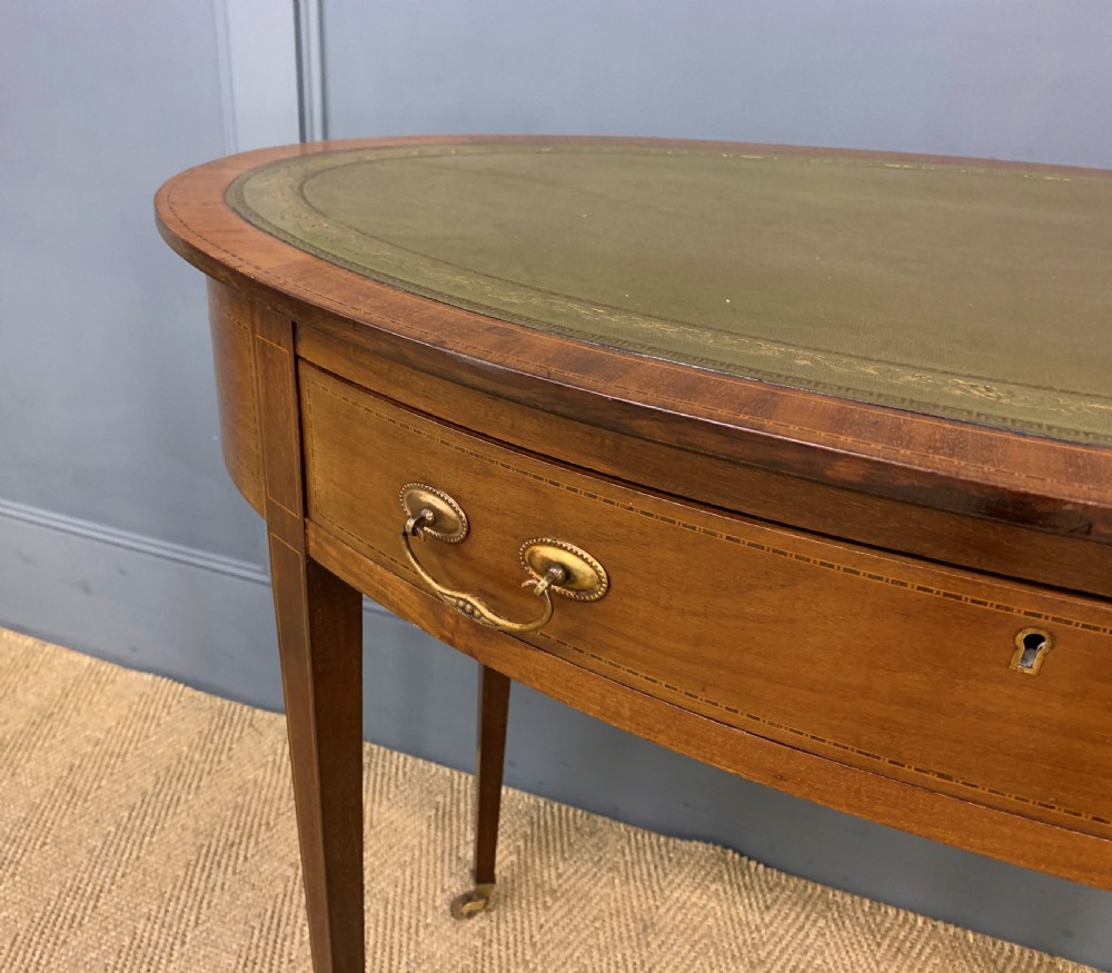An Edwardian oval mahogany writing table featuring a green leather inset top, single frieze drawer with brass bail handles, and square tapered legs with castors. View IV
