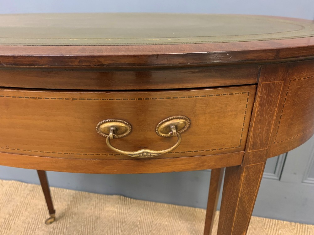 An Edwardian oval mahogany writing table featuring a green leather inset top, single frieze drawer with brass bail handles, and square tapered legs with castors. View V