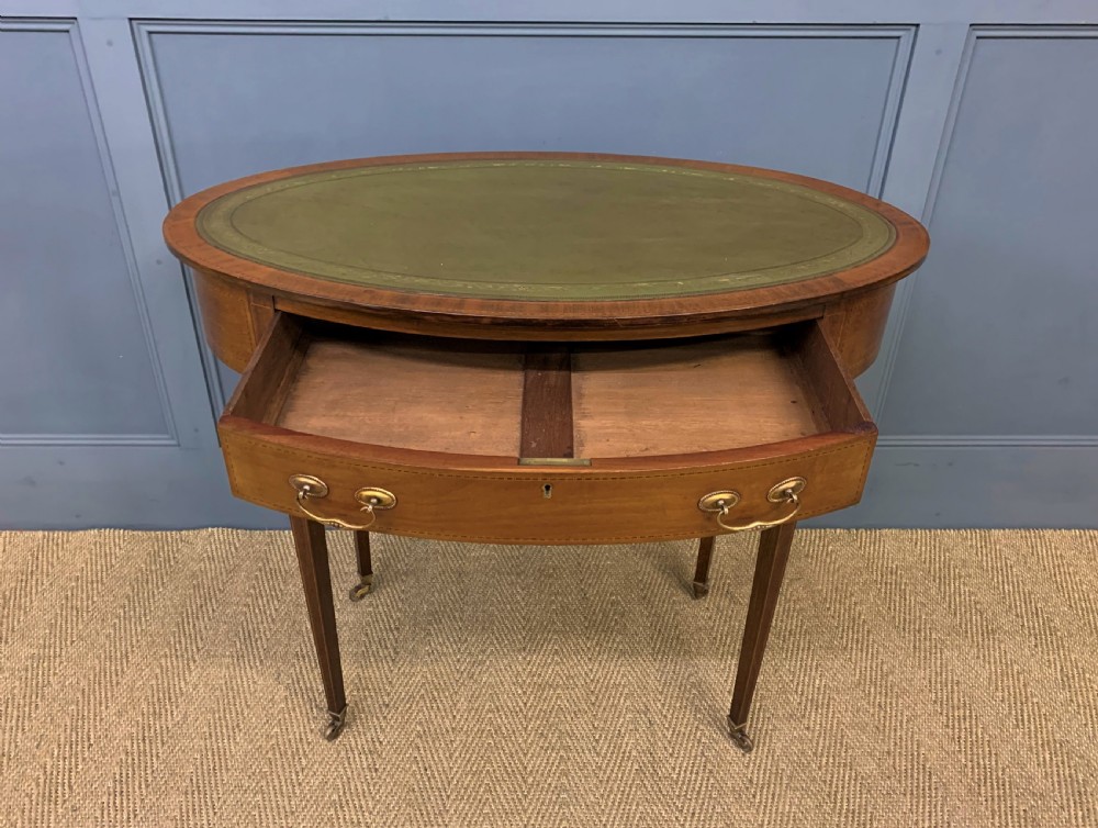 An Edwardian oval mahogany writing table featuring a green leather inset top, single frieze drawer with brass bail handles, and square tapered legs with castors. View VII